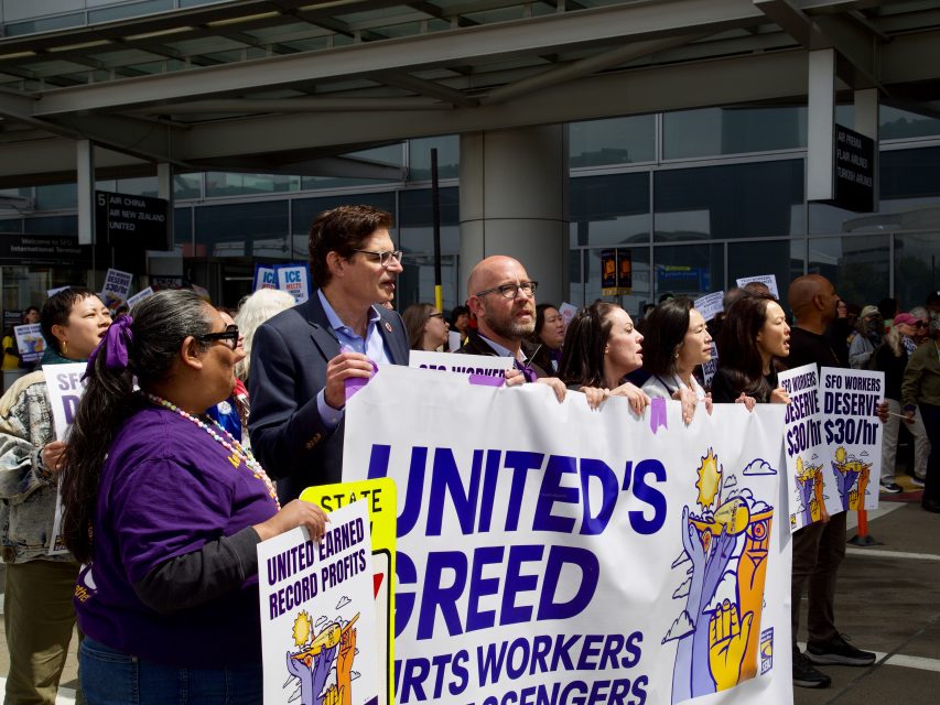 A group of people hold signs and a banner reading "United's Greed Hurts Workers & Passengers" during a protest outside an airport terminal, as police monitor the scene for any potential arrest.