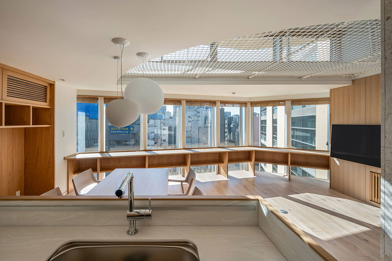 Modern open-plan room with large windows, built-in wooden seating, a wall-mounted TV, and a kitchen sink in the foreground, with cityscape views outside.