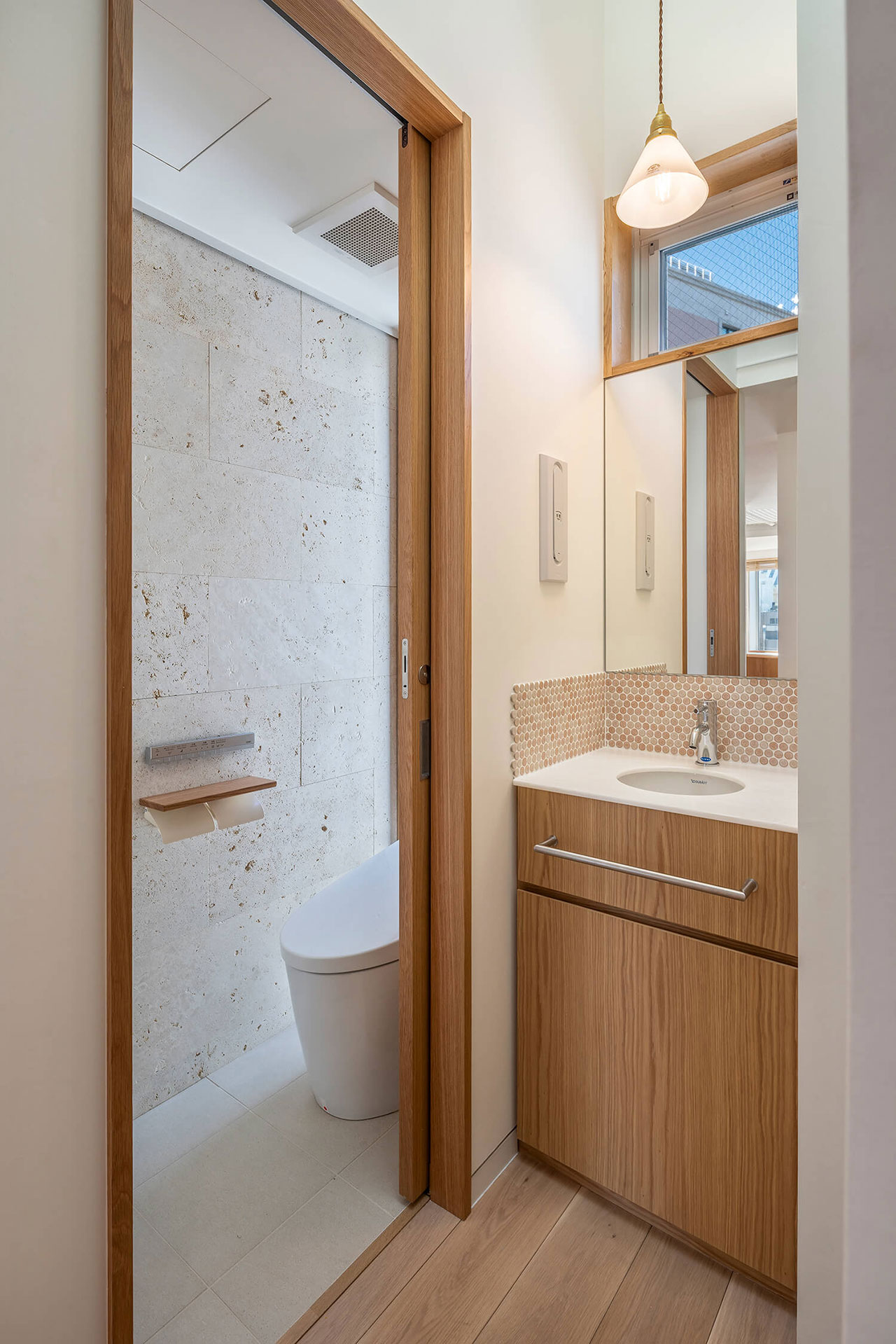 Modern bathroom with a wood vanity, sink, mirror, pendant light, and a separate toilet area with a sliding door and beige tiled walls.