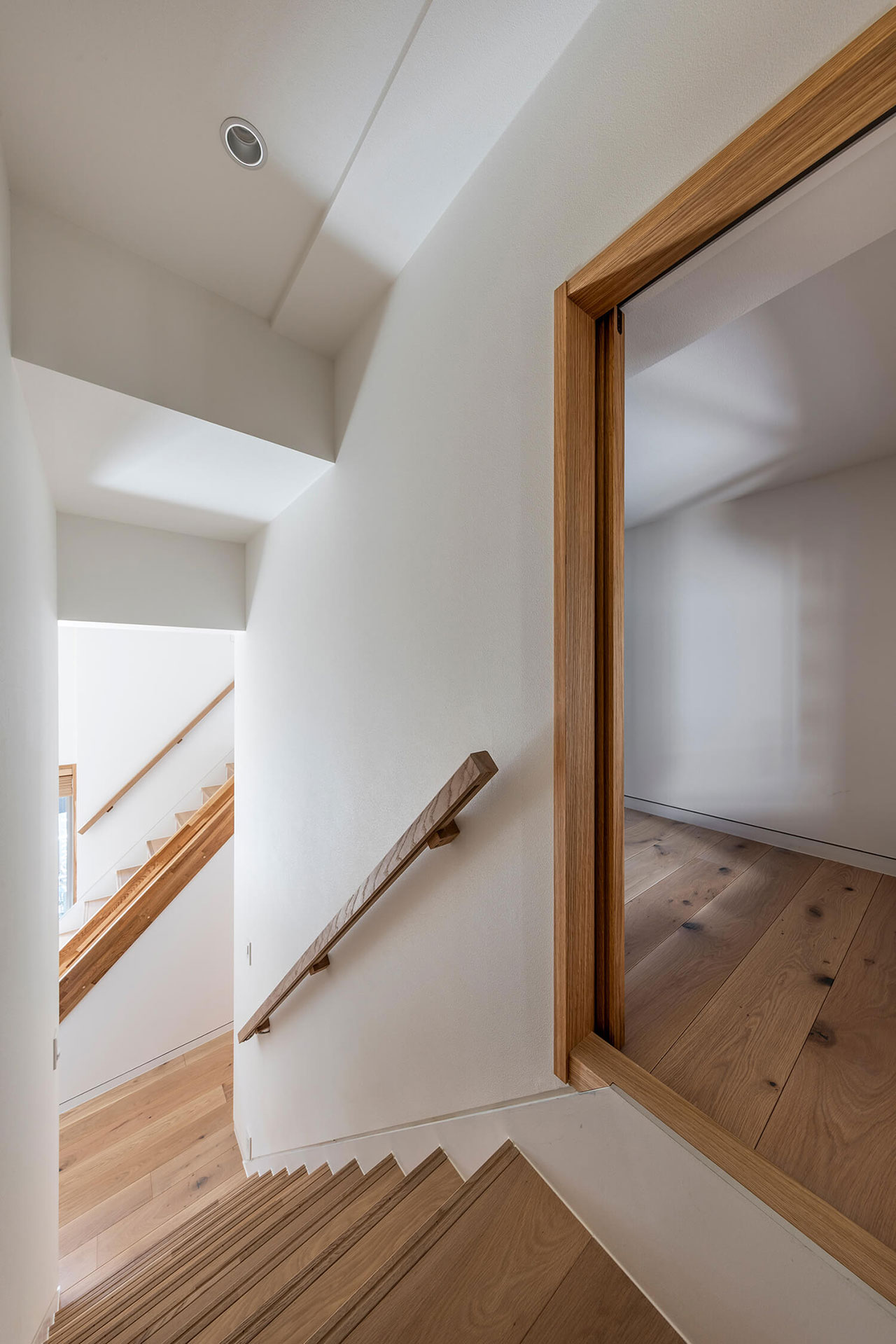 A modern indoor staircase with wooden steps and handrail, white walls, and an open doorway leading to a room with wooden flooring.