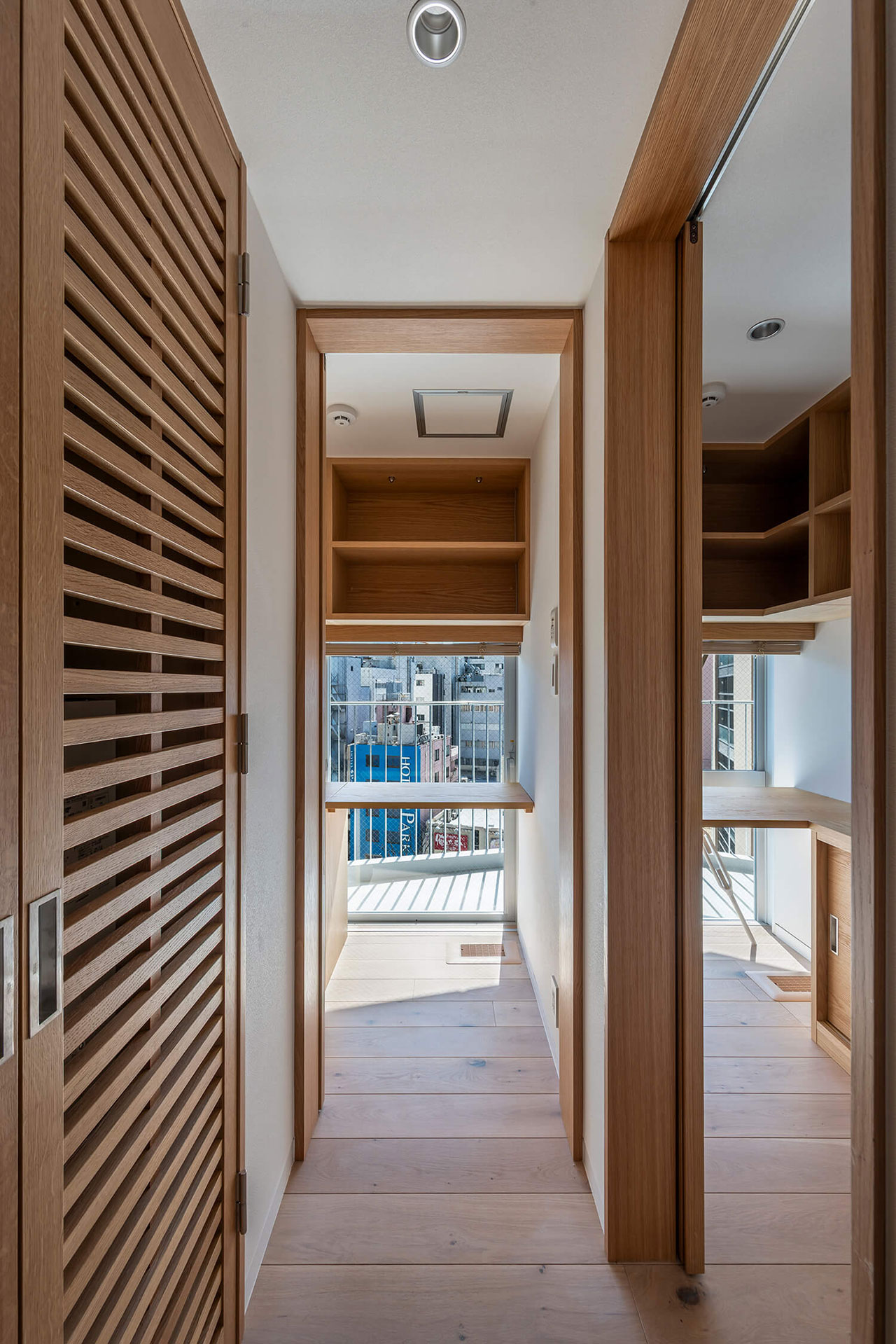 Narrow hallway with wooden flooring and built-in wooden shelves, leading to a window with a city view and a small desk area.