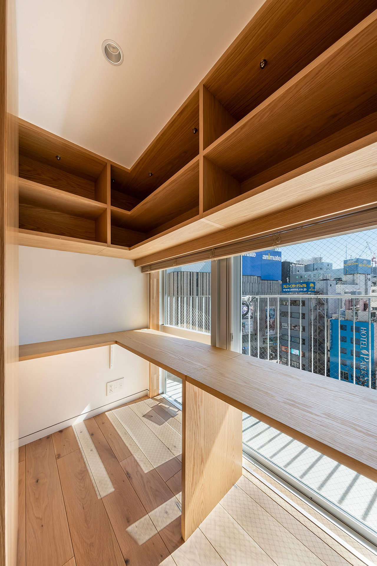 A small, modern workspace with wooden shelves, a built-in desk, large windows, and a city view in the background.