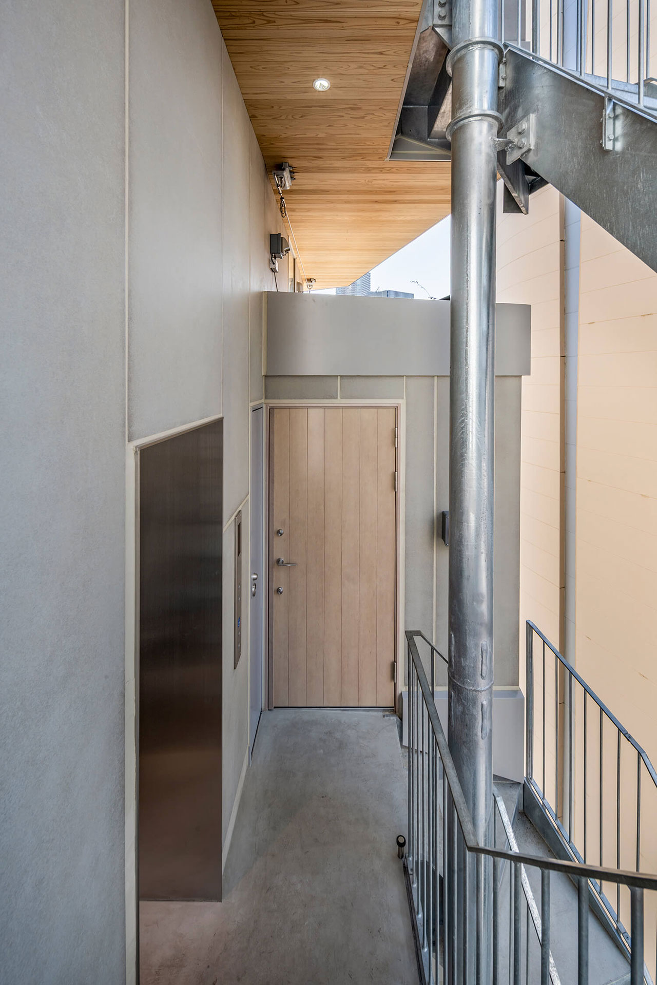 A narrow, modern apartment corridor with a wooden door, metal railing, exposed pipes, and concrete walls and floor under a wood-paneled ceiling.