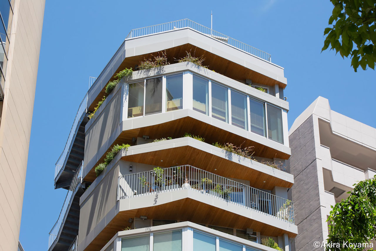 A modern multi-story building with angular balconies, large windows, and greenery on each level, set against a clear blue sky.