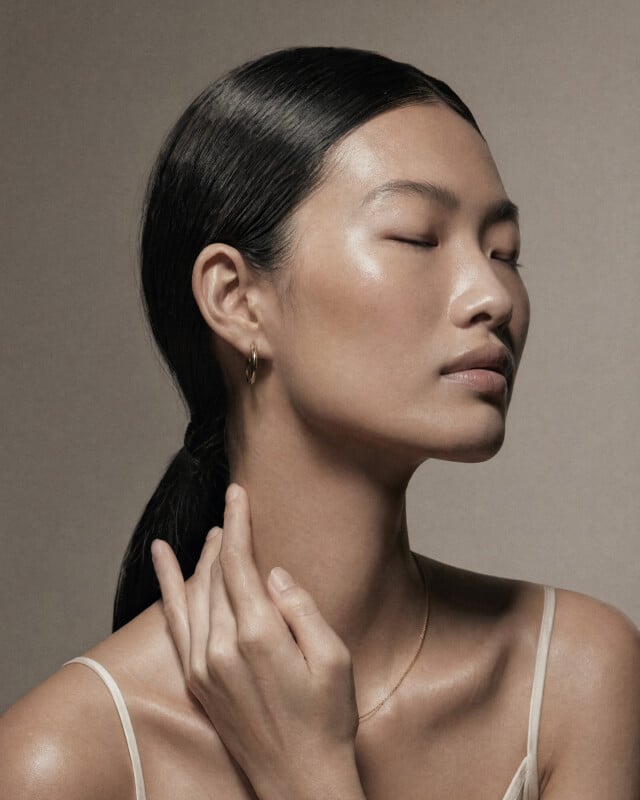 A woman with smooth, glowing skin and long, dark hair pulled back poses with her eyes closed, wearing gold hoop earrings and a thin necklace, her hand gently touching her neck against a neutral background.