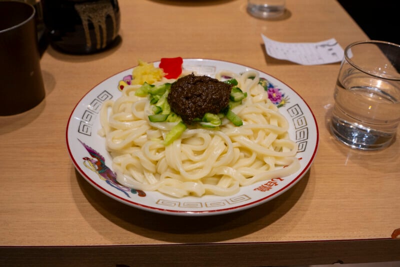 A plate of udon noodles topped with a dark sauce, sliced cucumber, grated ginger, and a red garnish sits on a wooden table next to a glass of water and a receipt.