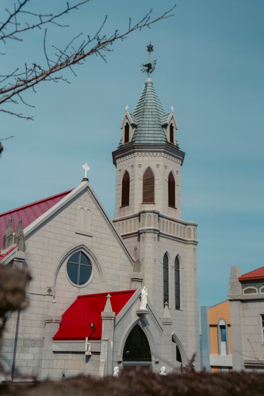 A stone church with a tall steeple and cross, red roofs, arched windows, and statues, set against a clear blue sky. Branches from a nearby tree are visible in the foreground.
