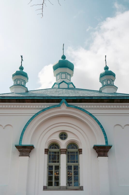 A white building with teal domes and arched windows, featuring ornate architectural details, set against a partly cloudy sky.