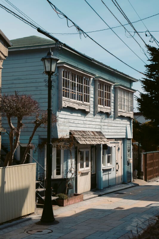A two-story light blue wooden house with large windows and a small entrance sits on a quiet, sunlit street. A vintage-style streetlamp stands nearby, and trimmed trees and fences line the sidewalk.