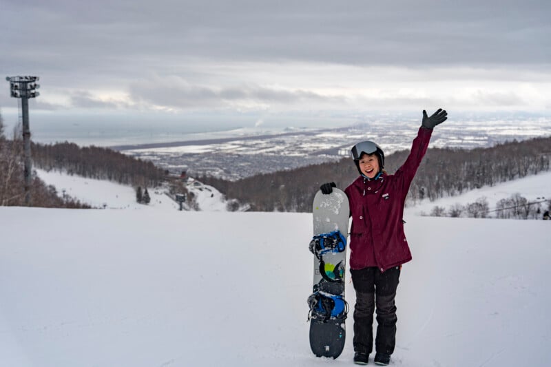 A person in a maroon jacket and black pants stands on a snowy slope, smiling with one arm raised and the other holding a snowboard upright. Snowy hills and a cloudy sky are visible in the background.
