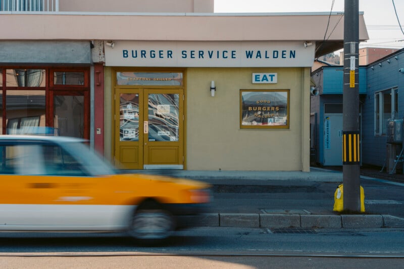 A yellow car drives past a restaurant called "Burger Service Walden" with yellow doors and a sign that says "EAT." The building has a beige exterior and what appears to be a burger sign in the window.