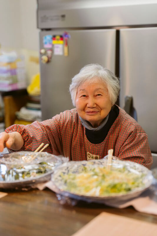 An elderly woman with gray hair smiles warmly while sitting at a table with plates of food covered in foil and chopsticks in front of her, in a cozy kitchen setting.