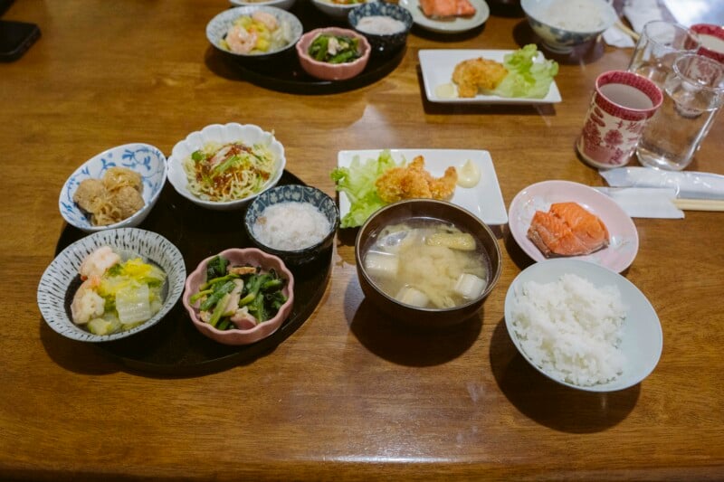 A Japanese meal set on a wooden table includes bowls of rice, miso soup, pickled vegetables, salad, fried food, a slice of salmon, and various small side dishes, with drinks and more plates in the background.