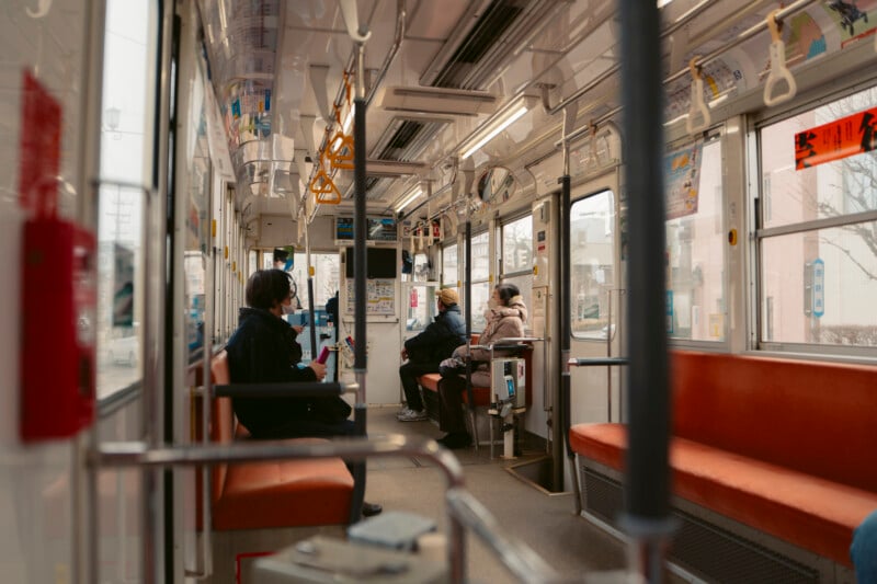 Several people sit quietly inside a public tram with orange seats, wearing winter clothing. Sunlight comes through large windows, and the cityscape is visible outside. The atmosphere is calm and reflective.