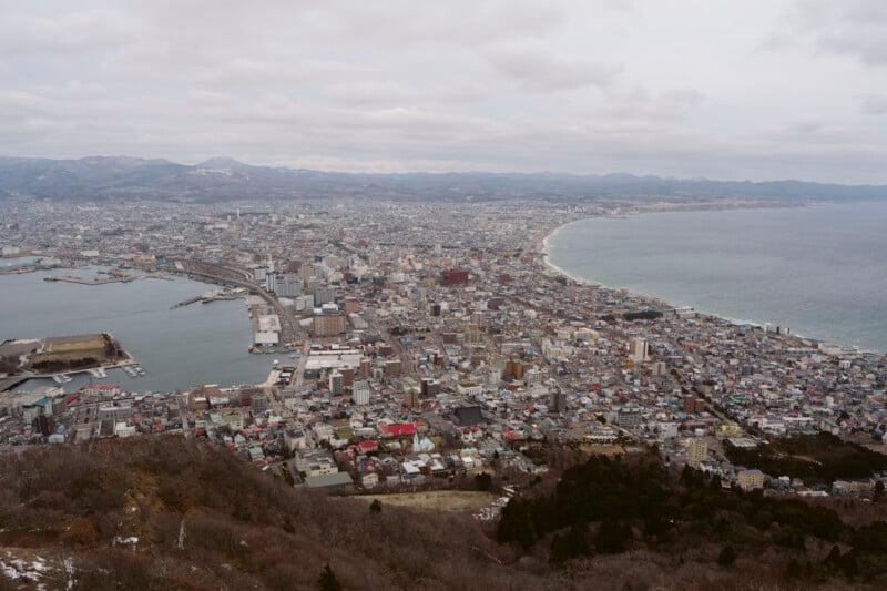 Aerial view of a coastal city with a curved shoreline, dense buildings, a harbor, and mountains in the distance under a cloudy sky.