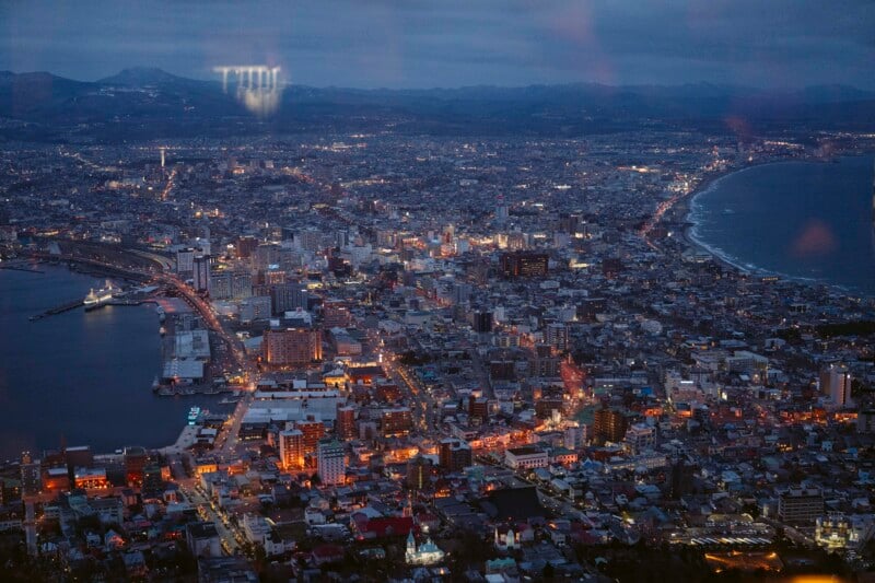 Aerial view of a coastal city at dusk, with buildings lit up, roads illuminated, a curved shoreline, and mountains in the background under a darkening sky.