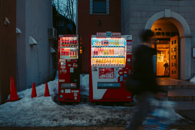 Two red vending machines stand outside a building at dusk, surrounded by snow and orange cones. A blurred person walks past, holding a bag, while warm light glows from the building's entrance in the background.