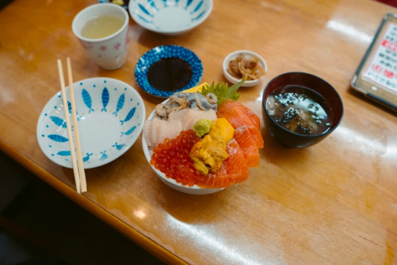 A bowl of assorted sashimi with rice, including salmon, sea urchin, and roe, sits on a wooden table next to empty plates, chopsticks, a cup of tea, miso soup, and small side dishes.