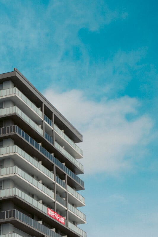 A modern apartment building with multiple balconies stands against a blue sky. A red sign with white Japanese text hangs on one of the lower floors.