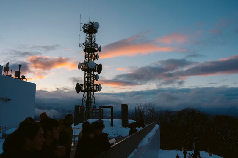 A group of people stands on a snowy rooftop at sunset, next to a large telecommunications tower with satellite dishes and antennas. The sky is partly cloudy with pink and orange hues.