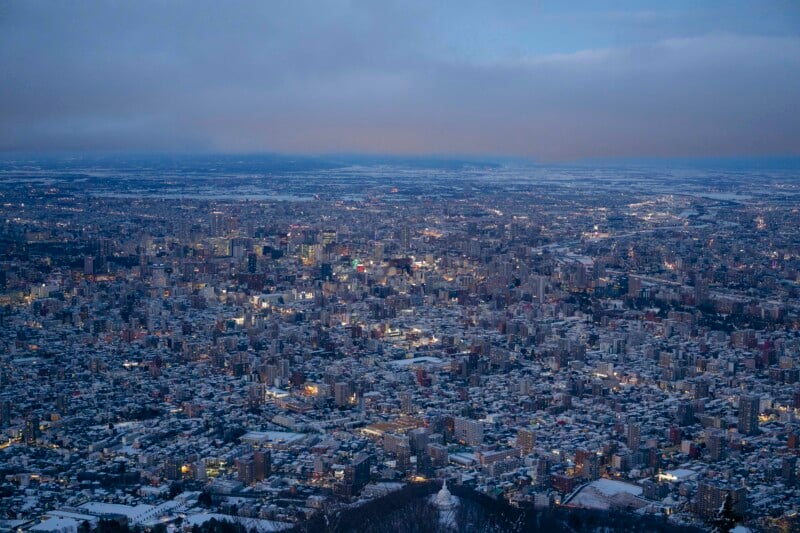 Aerial view of a sprawling cityscape at dusk, with buildings and streets dusted in snow. City lights are glowing, and the sky is cloudy with a faint light on the horizon.
