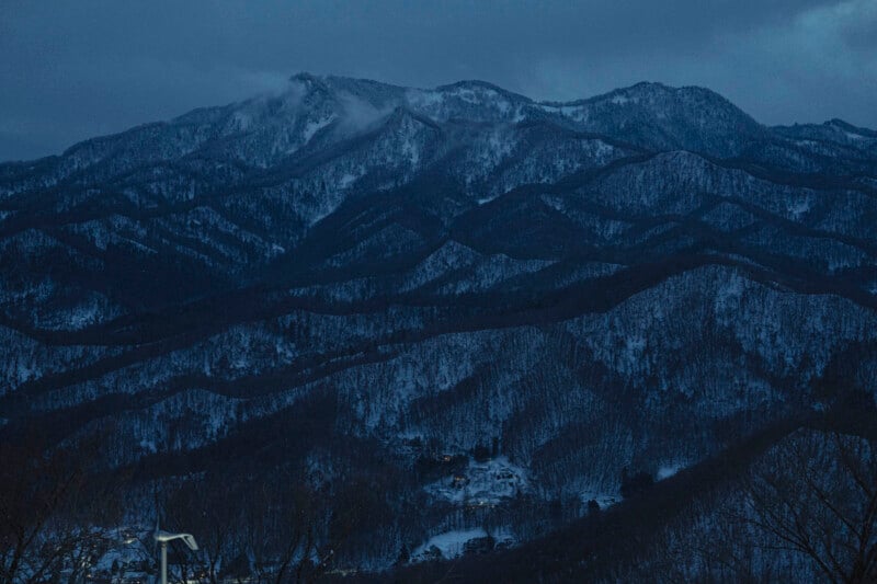 Snow-covered mountains under a cloudy, dusky sky. The slopes are densely forested with leafless trees, and a few small buildings with lights can be seen at the base of the mountains.
