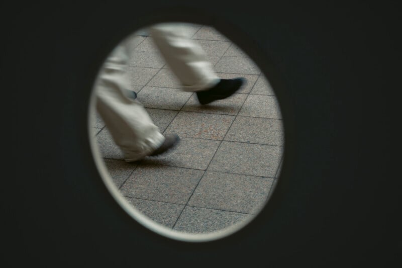 A pair of legs wearing light-colored pants and dark shoes are visible walking on a tiled floor, seen through a circular window or opening in a dark surface.
