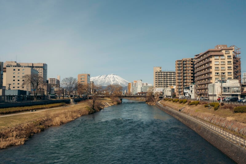 A river flows through a city with mid-rise buildings on both sides. In the background, a snow-capped mountain rises under a clear blue sky. Trees line the riverbanks, and a bridge spans the water further ahead.