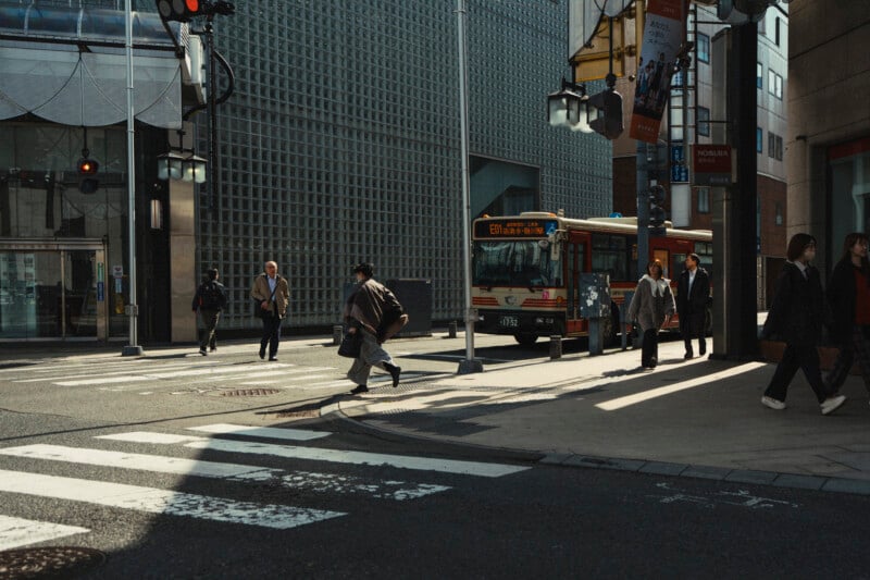 People cross a city street at a crosswalk near a bus, with modern buildings and traffic lights in the background. The scene is lit by soft daylight and features a mix of pedestrians, some in motion and some standing.