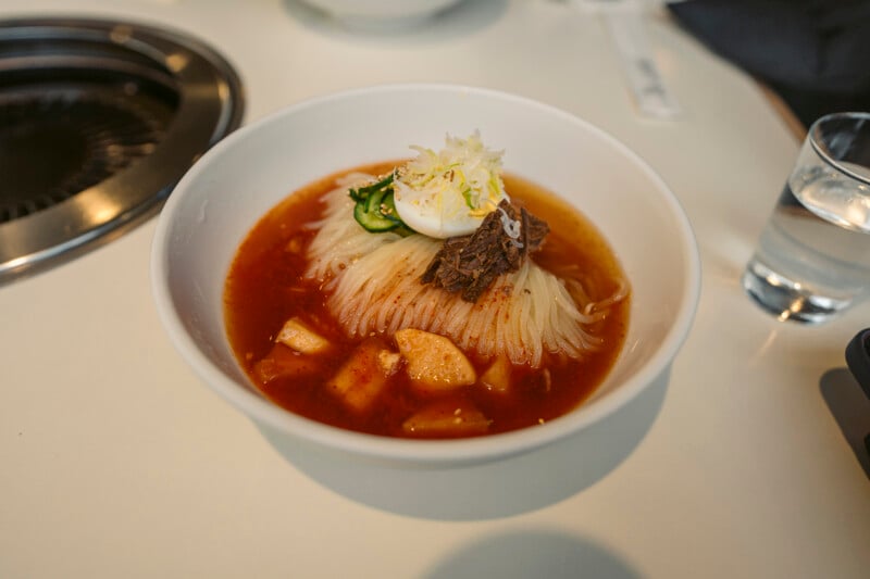 A bowl of Korean cold noodles in red broth topped with sliced cucumber, beef, a boiled egg half, and shredded vegetables, sits on a white table next to a glass of water.