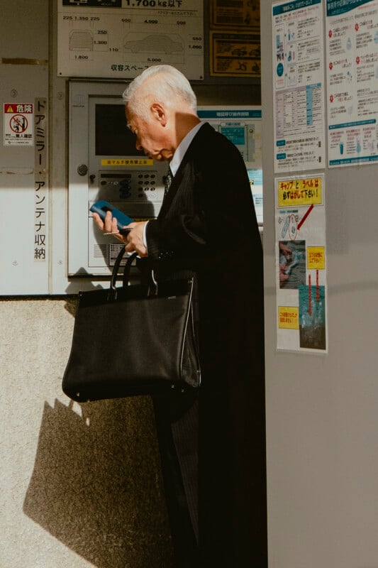 An older man in a suit stands near a ticket or payment machine, holding his phone and a large black briefcase. Sunlight casts shadows on him and the wall, which is covered with various informational posters in Japanese.