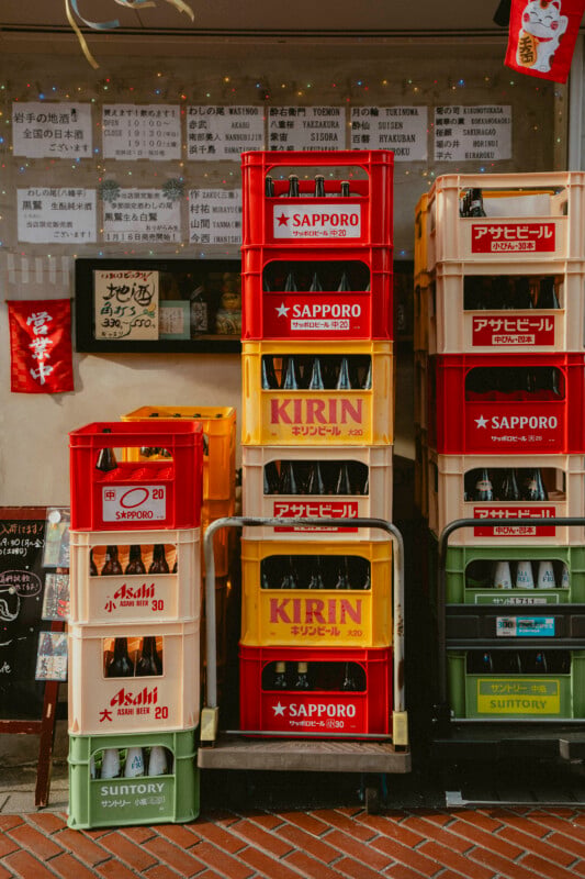 Plastic crates labeled Sapporo, Kirin, Asahi, and Suntory are stacked outside a Japanese restaurant, with drink bottles inside and handwritten menus displayed on the wall behind them.