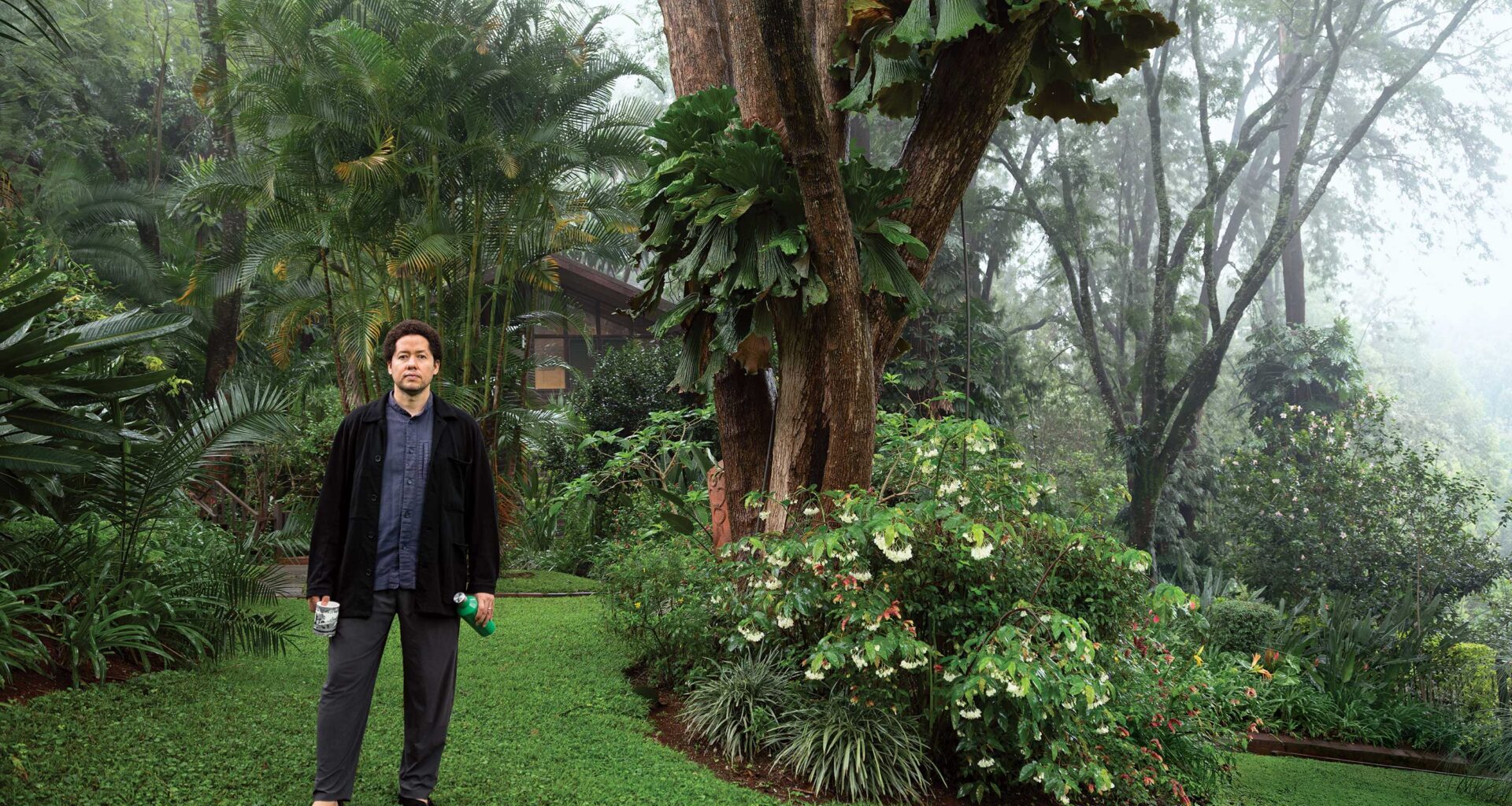 Man in a garden holding a water bottle, surrounded by lush green plants and trees on a misty day.