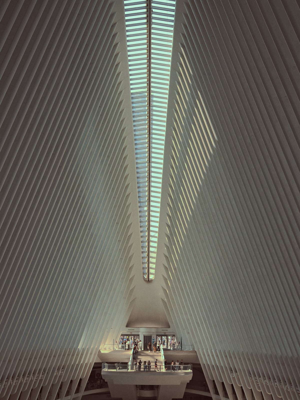 Interior view of a modern architectural building with tall, symmetrical white ribs and a skylight above. People are gathered on a balcony at the far end, illuminated by natural light.