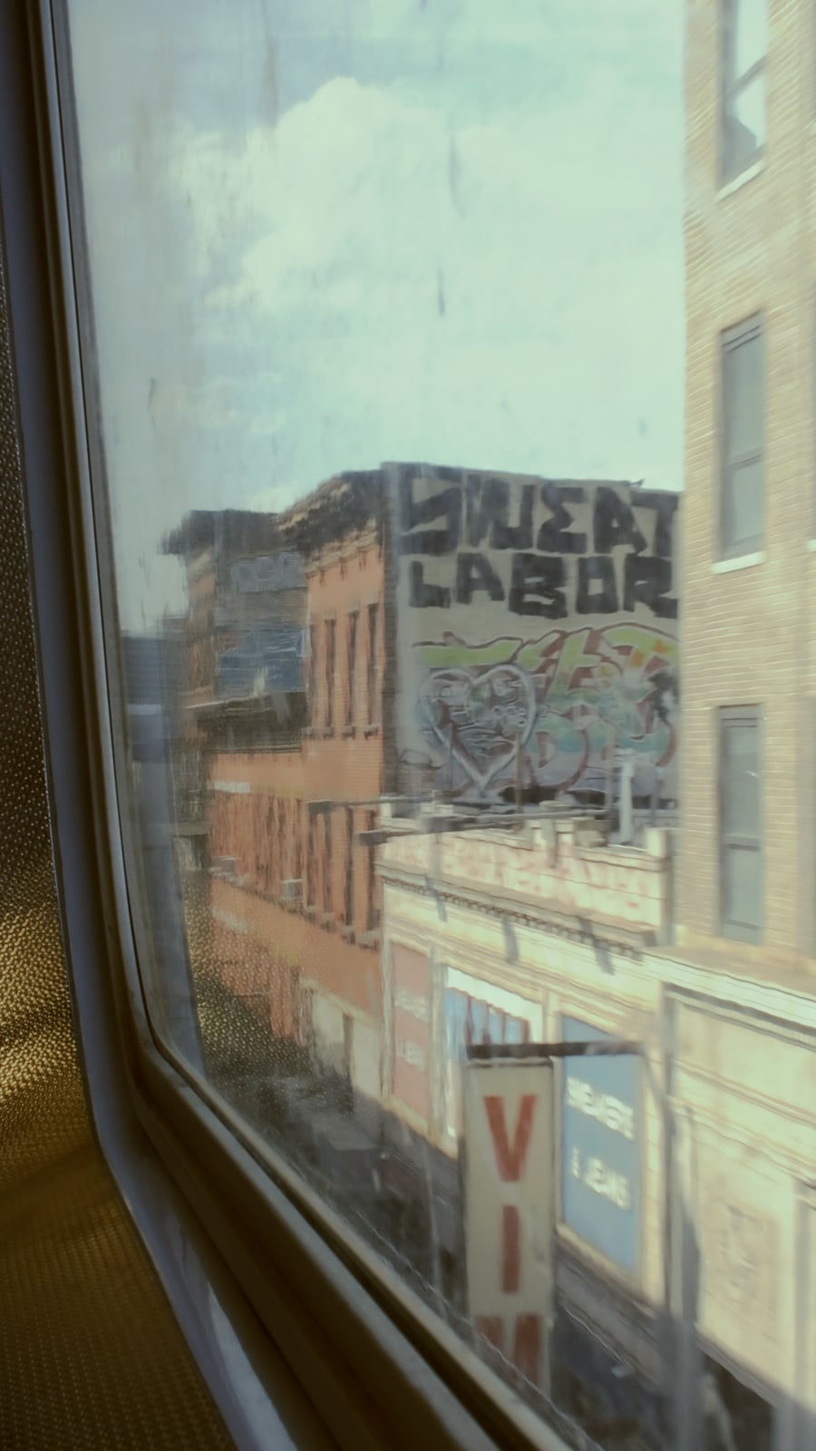 View through a window showing a city street with brick buildings, graffiti art on a wall reading "CHEAP LABOR," and a partially visible sign that says "VIA." The scene is slightly blurred.
