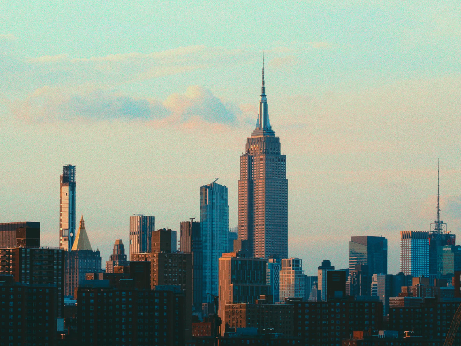 A city skyline at sunset featuring the Empire State Building and surrounding skyscrapers under a partly cloudy sky.