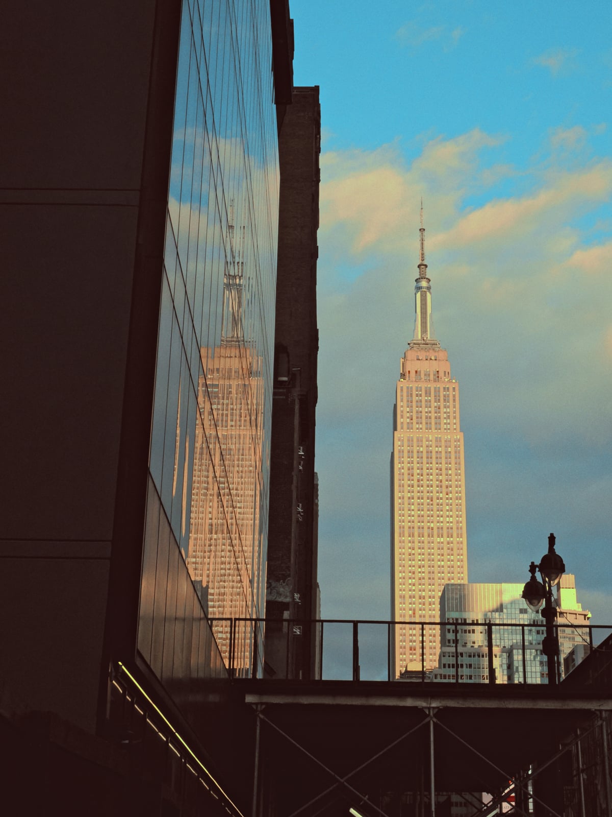 The Empire State Building stands tall against a blue sky with clouds, its reflection visible on the glass windows of a nearby building in the foreground.