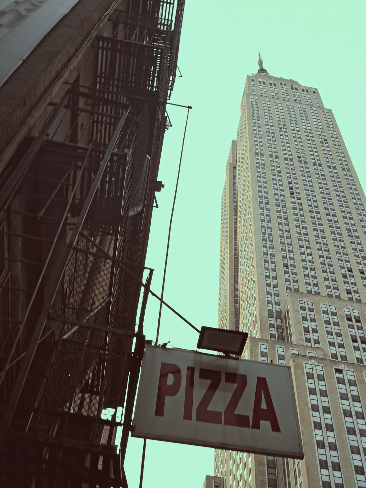 A tall city building towers into the sky next to a fire escape, with a large sign in the foreground reading "PIZZA" in red letters. The photo is taken from a low angle, looking upward.