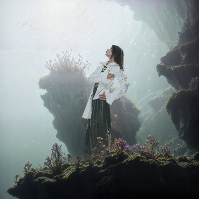A woman in a white jacket and dark skirt stands on mossy ground, surrounded by colorful plants, looking upward toward a glowing, ethereal light above rocky cliffs. The scene feels surreal and otherworldly.