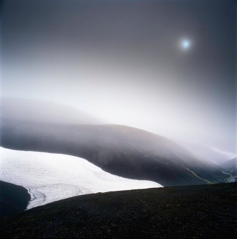 A misty, desolate landscape with rolling dark hills, patches of lingering snow, and a hazy sun barely visible through thick fog in the sky. The scene appears cold, remote, and tranquil.