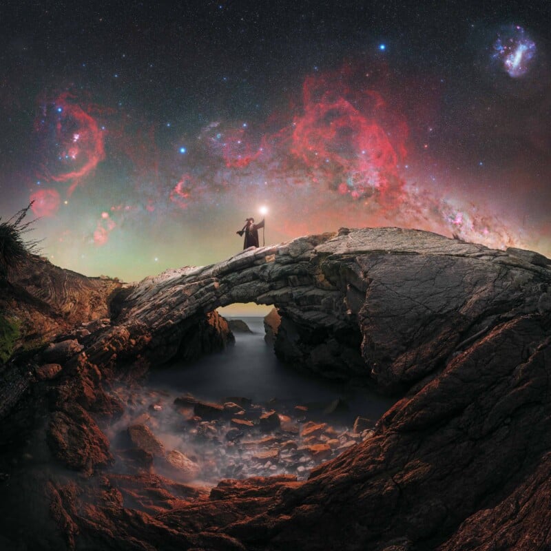 A person stands on a rocky arch holding a glowing staff, overlooking a misty shore with a vibrant, colorful night sky filled with stars and nebulae above.