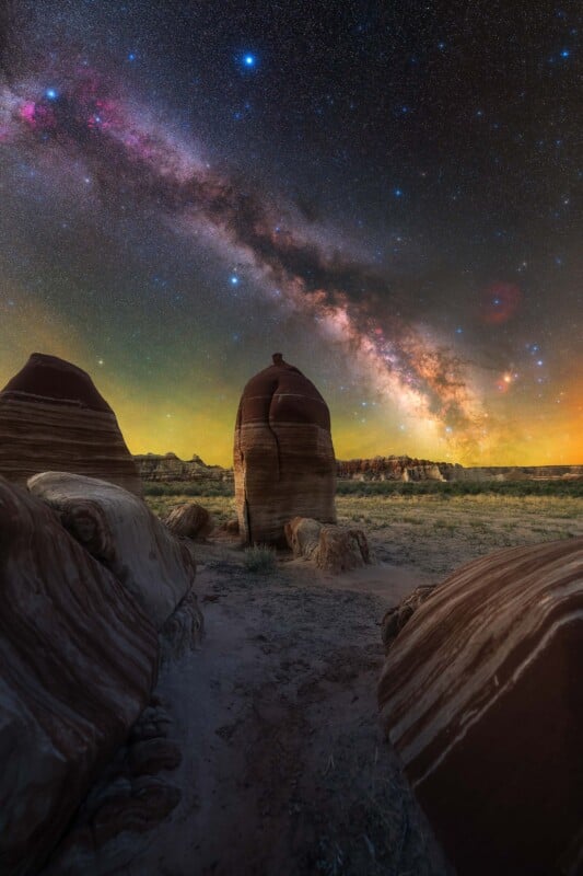 A dramatic view of striped rock formations under a vivid, star-filled night sky with the Milky Way brightly arching overhead, casting colorful light across the desert landscape.