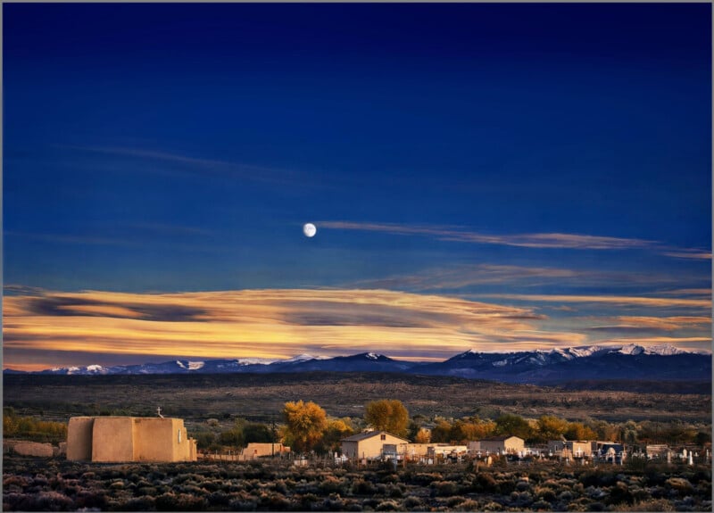 A serene landscape at dusk features adobe buildings, autumn trees, and mountains with snow-capped peaks under a deep blue sky with a bright moon and streaks of sunset clouds.
