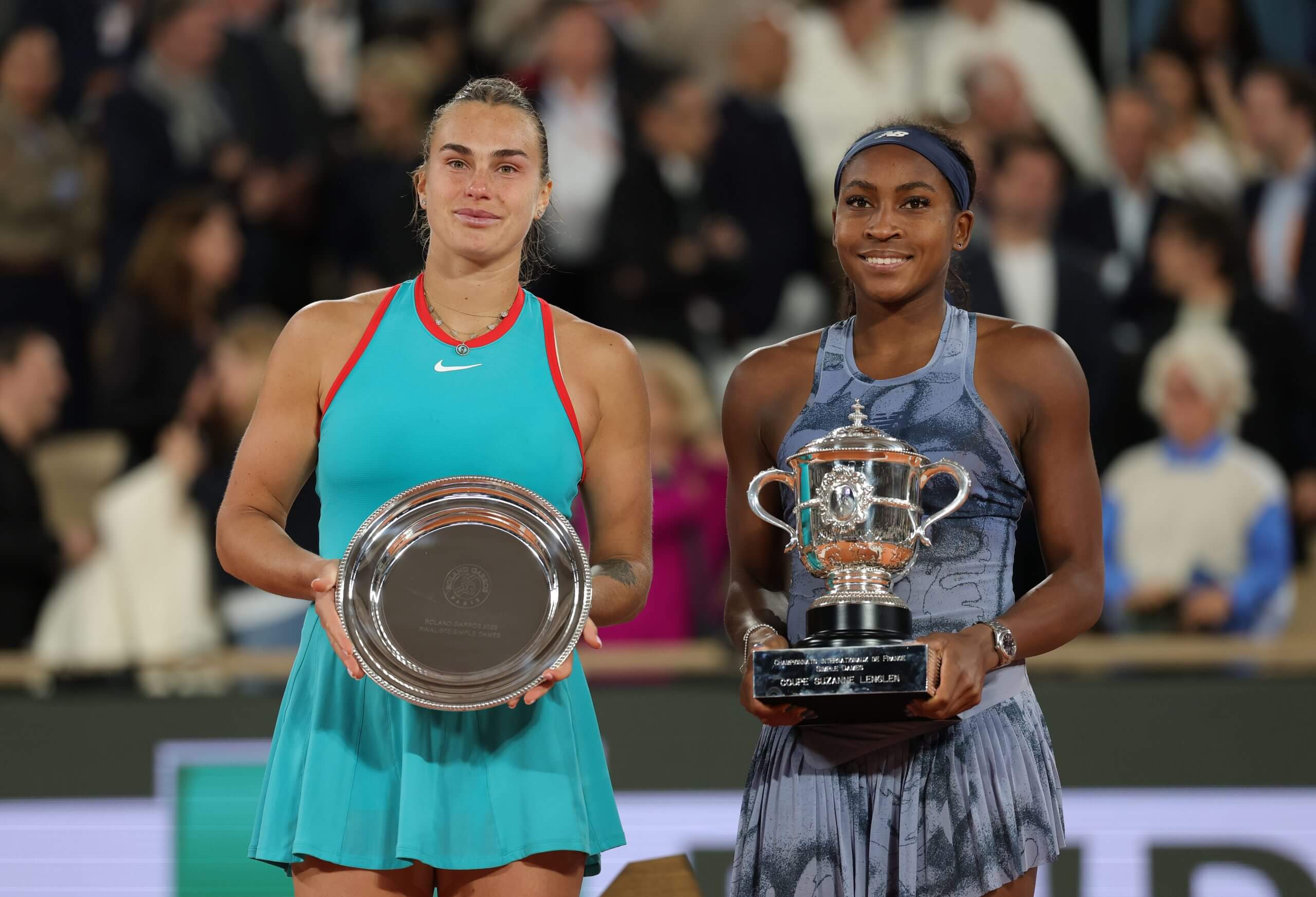 Aryna Sabalenka (left) holds a silver plate while standing next to Coco Gauff (right) who is holding a silver trophy.