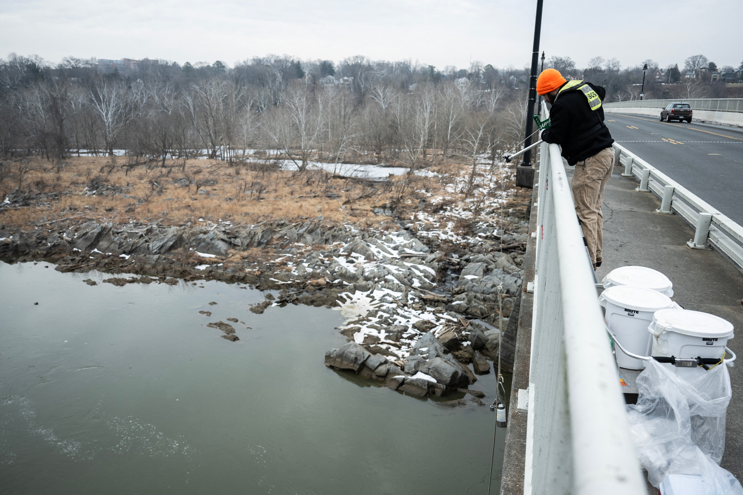 A man standing on a bridge lowers a rod into a waterway below. 