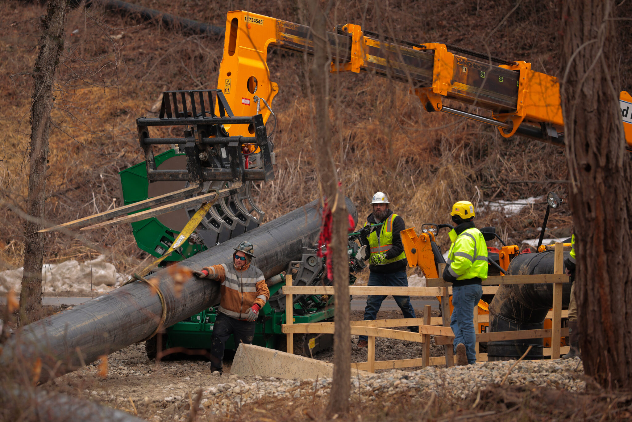 Three construction workers examine a large metal pipe with heavy machinery in the background. 