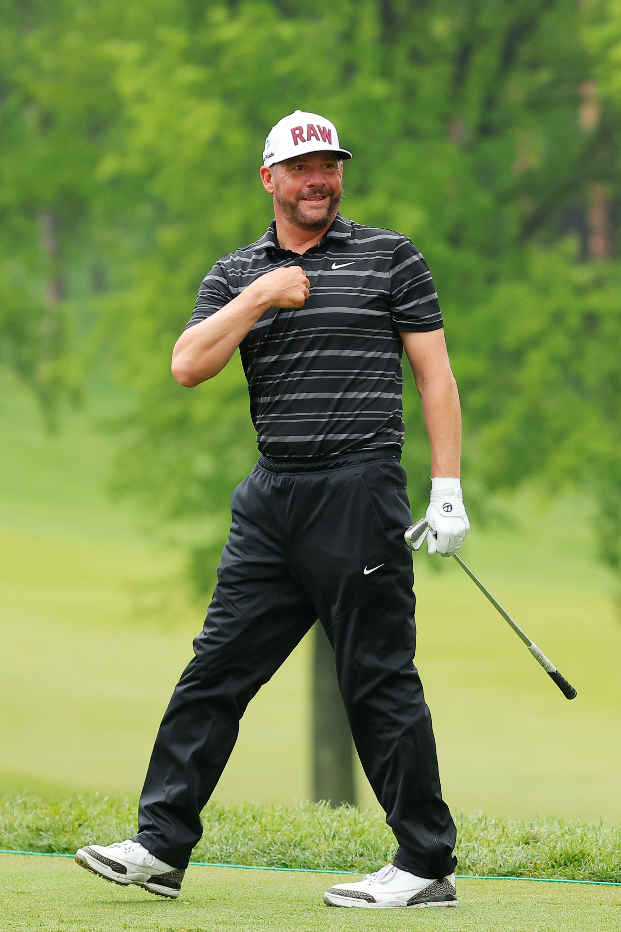 Golfer in black striped shirt and white cap walks on course, hand to chest.