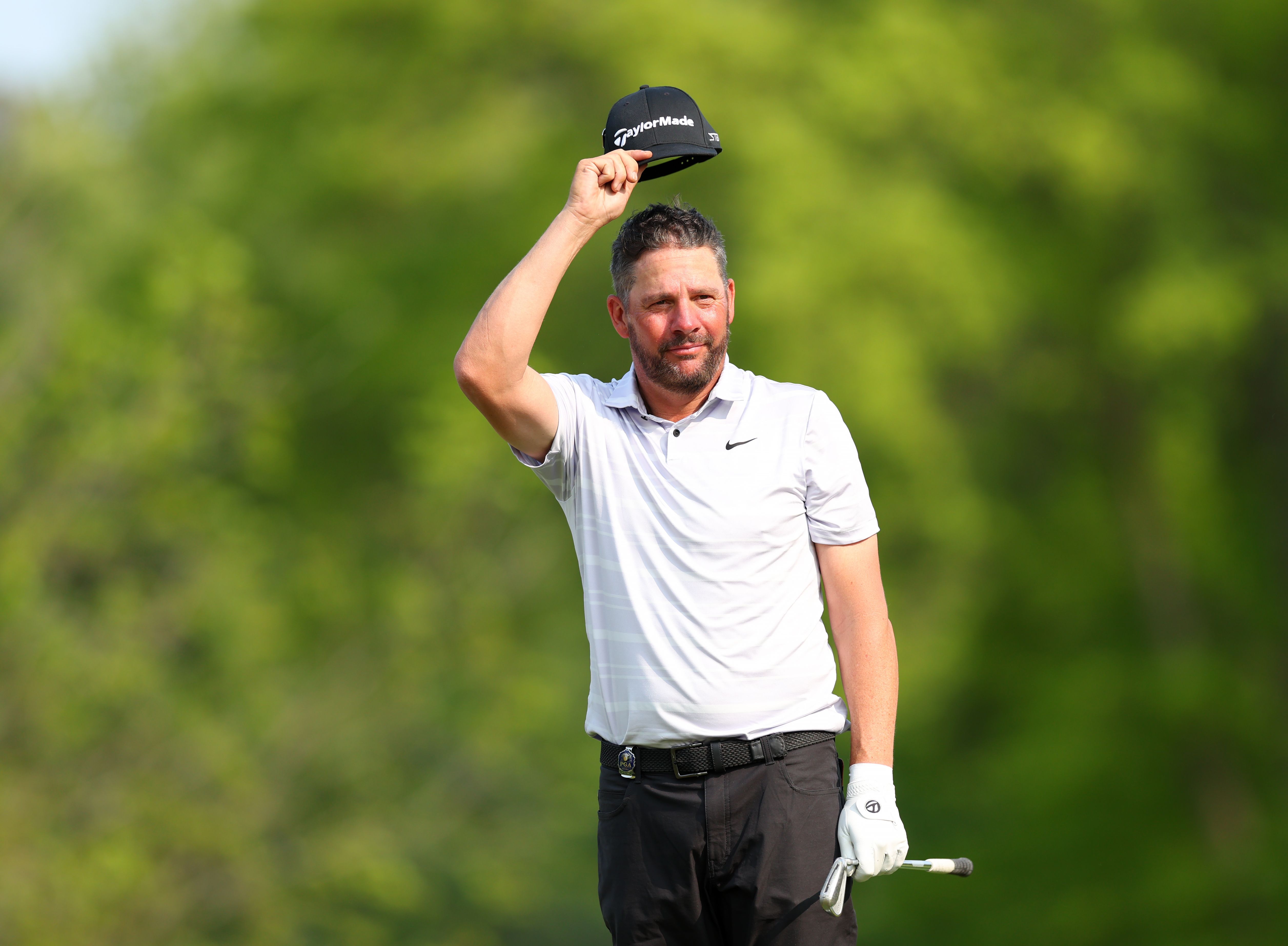 A male golfer in a white shirt raises his dark cap, acknowledging the crowd on a golf course.