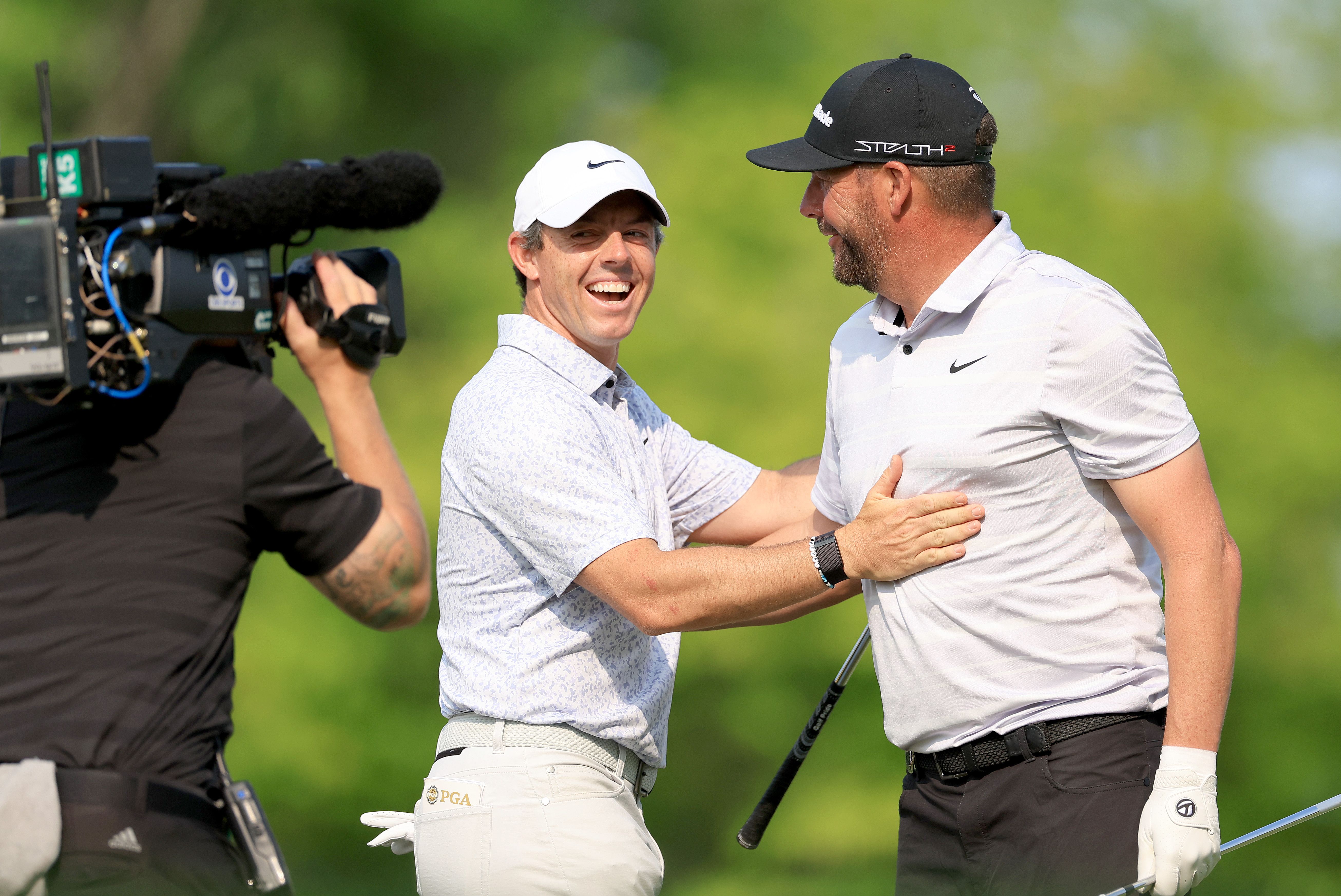 Rory McIlroy and Keegan Bradley shake hands on a green golf course near a camera.
