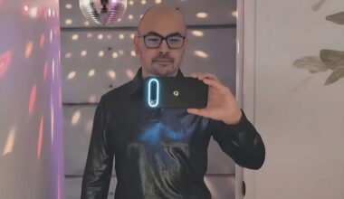 A person wearing a black shirt takes a selfie in a room with a disco ball and colorful light reflections, holding a smartphone with a glowing camera module.